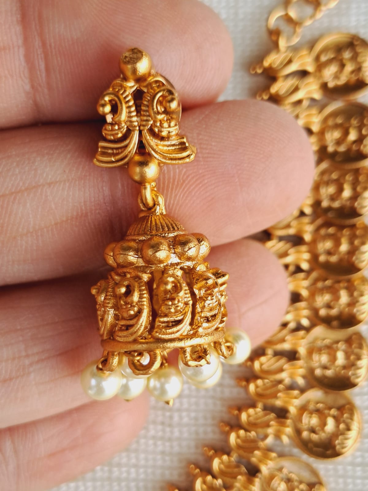 Intricate gold necklace with ornate pendant featuring delicate floral design and pearl accents, showcased in a hand-held display against a neutral background.