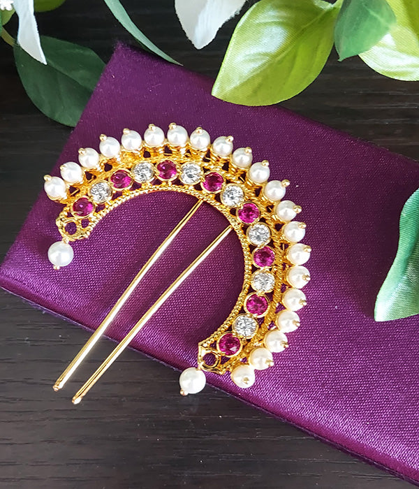 Ornate gold-toned hair accessory adorned with sparkling red gems and white pearls, resting on a vibrant purple fabric with lush greenery in the background.