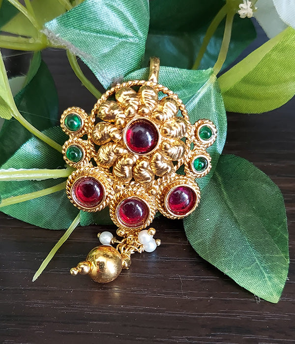 Ornate gold hair accessory with vibrant red and green gemstones, showcased on a wooden surface surrounded by lush green foliage.