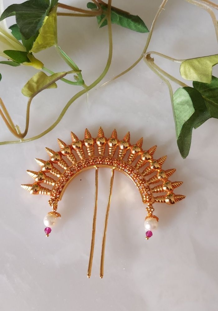 Ornate golden hair accessory with spiky design and ruby accents, displayed against a background of green leaves.