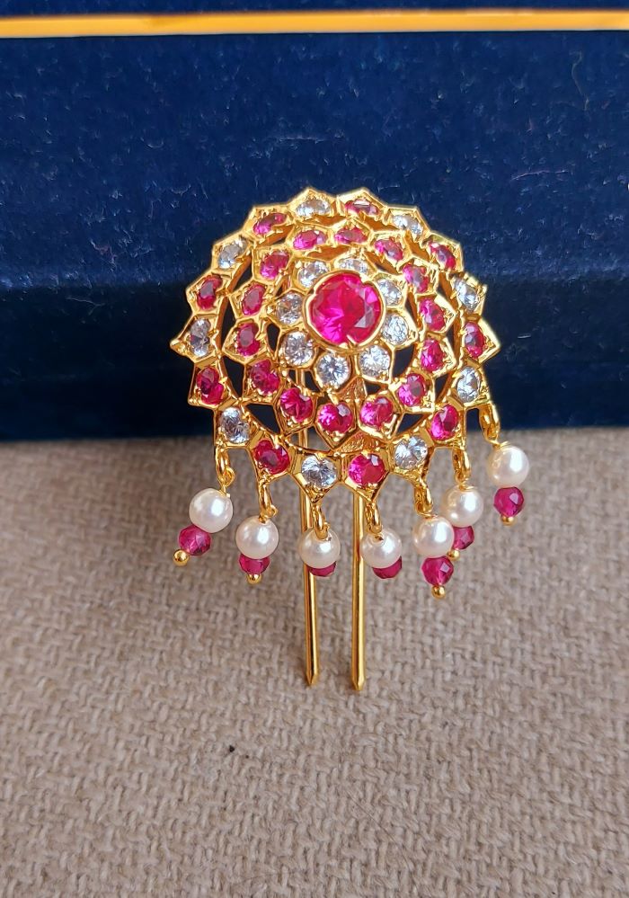 Ornate gold-toned floral hair accessory with vibrant pink gemstones and pearl accents, displayed on a dark blue background.