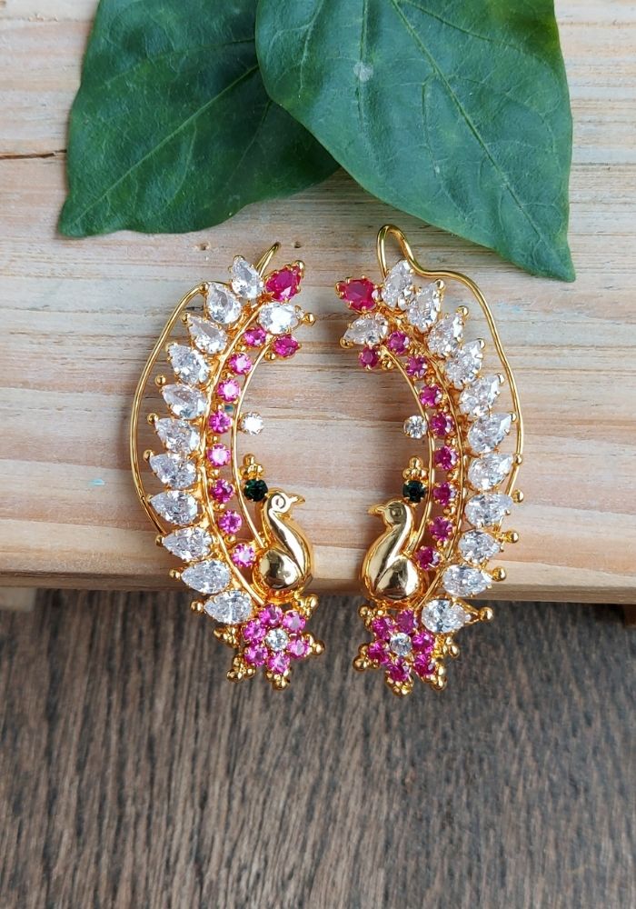 Ornate gold-toned hoop earrings with vibrant pink and clear crystal stones, set against a backdrop of lush green leaves on a wooden surface.