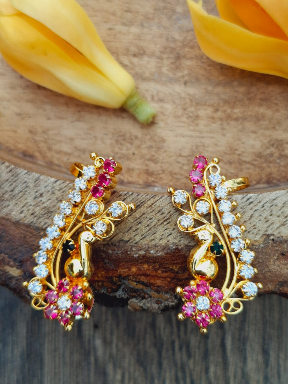 Vibrant gold-tone earrings adorned with pink and clear crystals in a floral design, placed on a wooden surface with yellow flowers in the background.