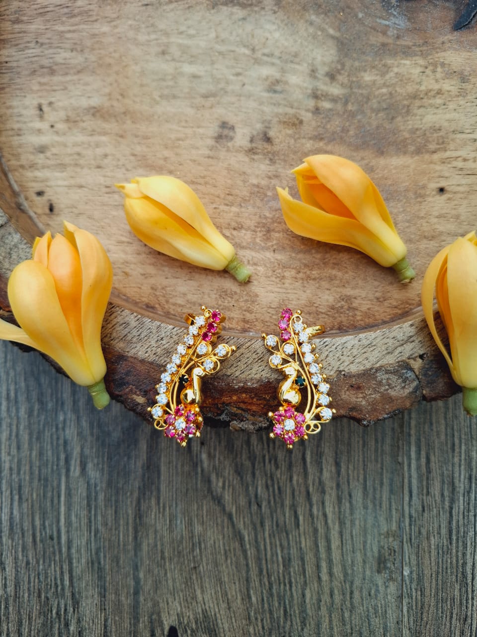 Ornate golden peacock-shaped earrings with vibrant red and pink gemstones, displayed on a rustic wooden surface with yellow flower petals.
