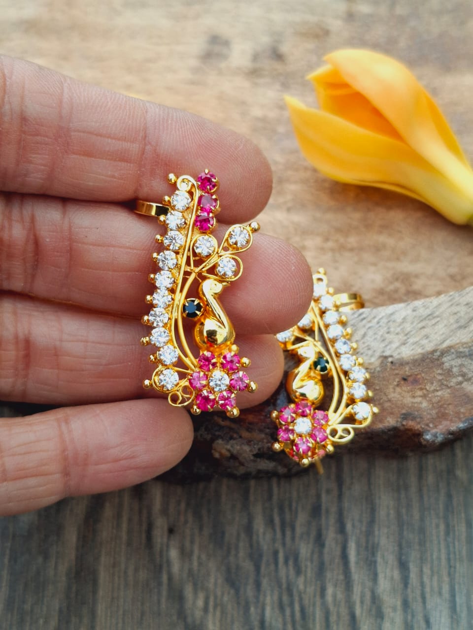 Intricate gold-toned earrings with sparkling red and white gems, showcased on a wooden surface with a yellow flower petal in the background.