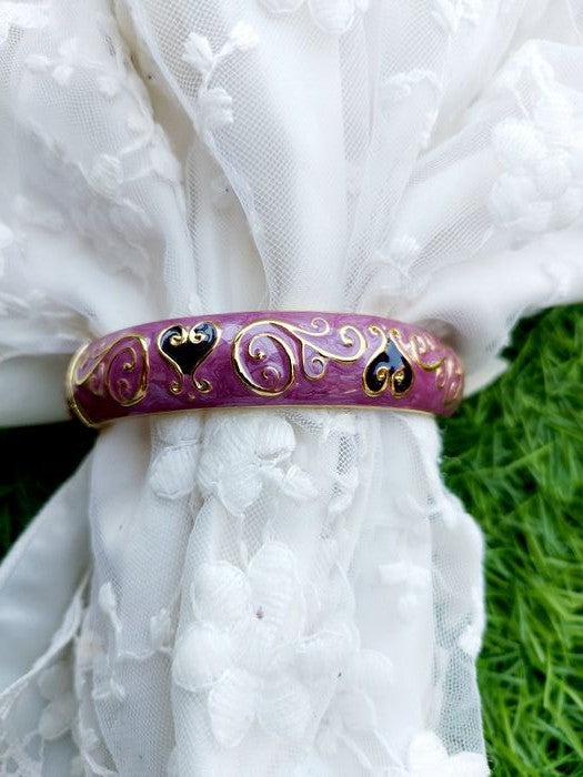 Elegant floral bangle bracelet in vibrant purple hue with intricate metal detailing, displayed against a backdrop of delicate white fabric and lush green grass.