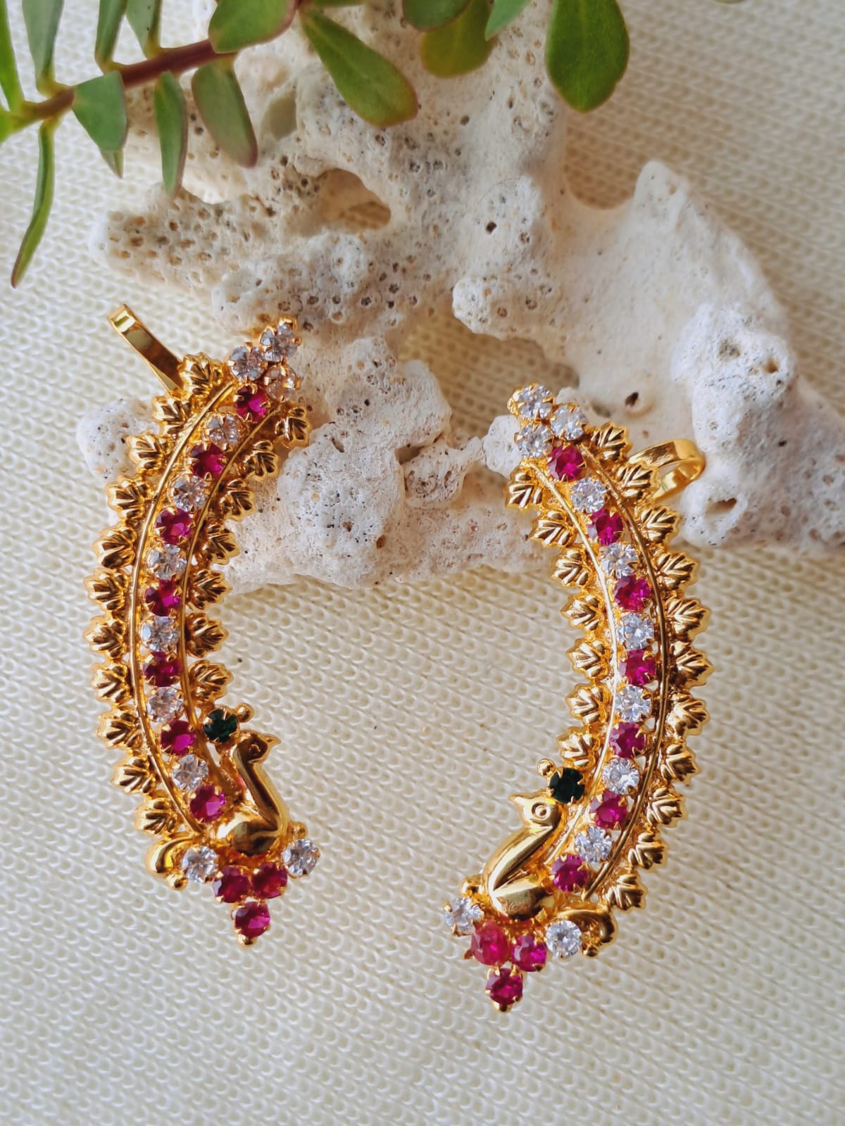 Elegant golden peacock-inspired earrings adorned with sparkling ruby-colored gemstones, set against a natural backdrop of leaves and crystals.