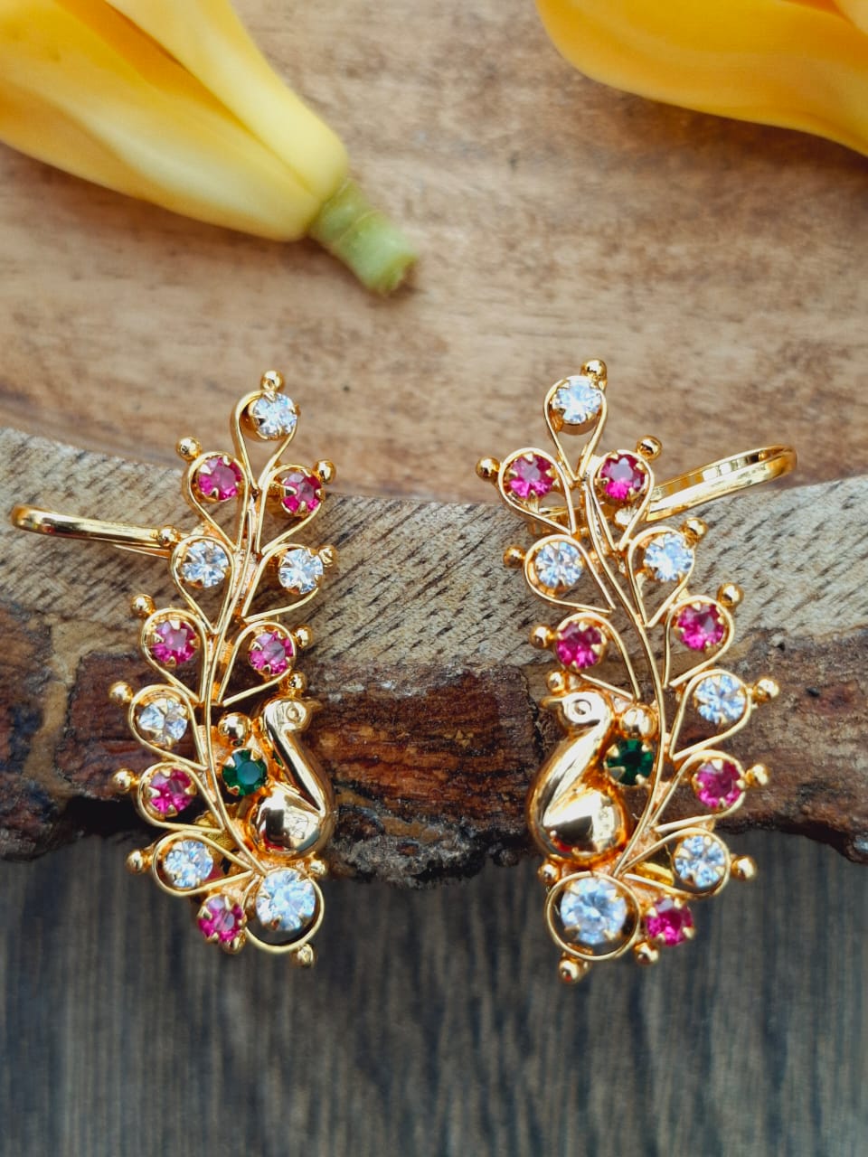Elegant golden peacock-inspired earrings adorned with colorful crystal gems, displayed on a rustic wooden surface.