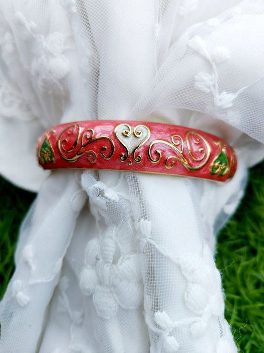 Vibrant Pink Kada
Shiny red metal bracelet with intricate floral designs, set against a backdrop of delicate white fabric and lush green foliage.