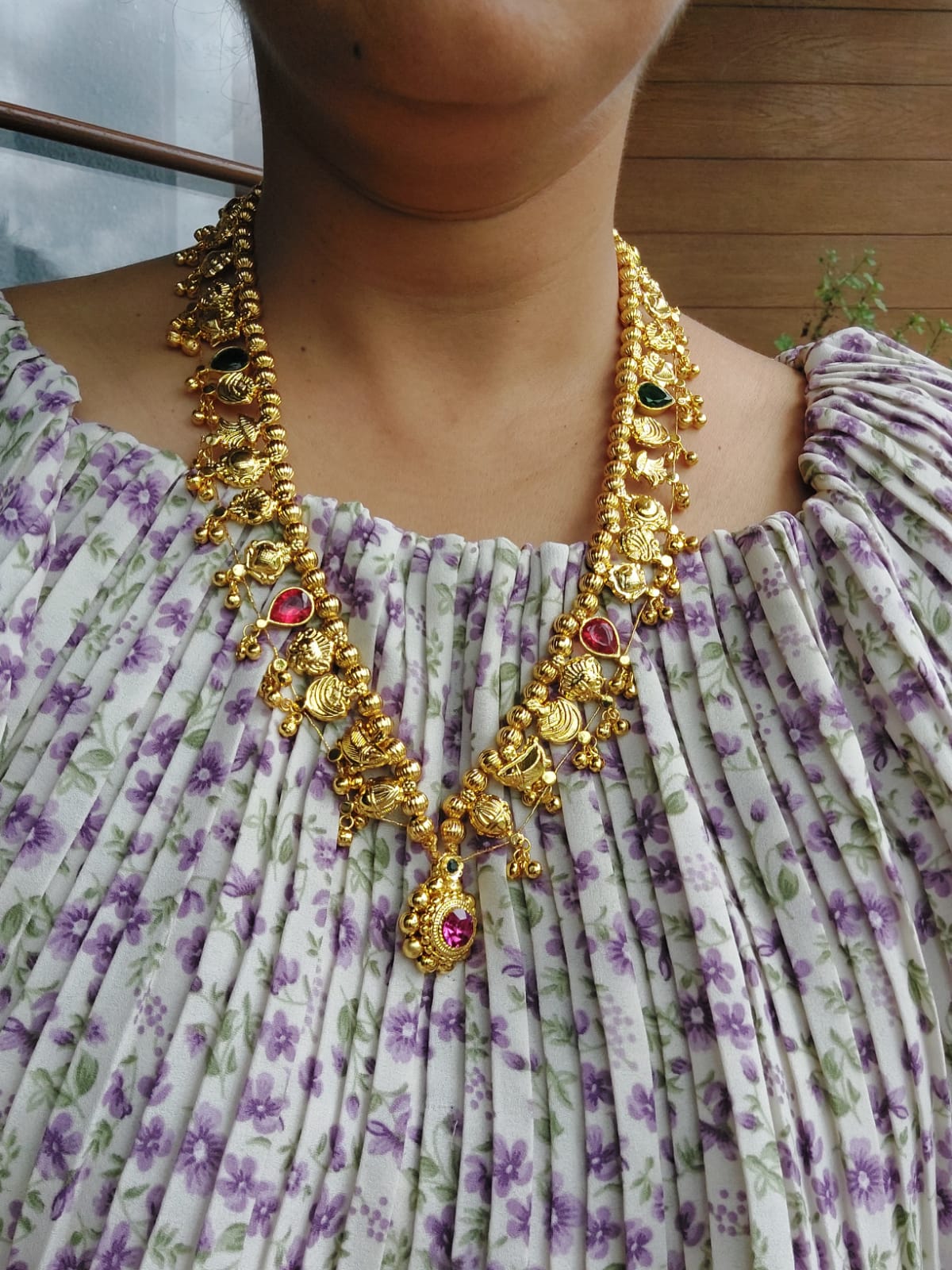 Ornate golden necklace featuring multicolored beads, decorative pendants, and intricate metalwork, worn by a person with a floral patterned dress in the image.