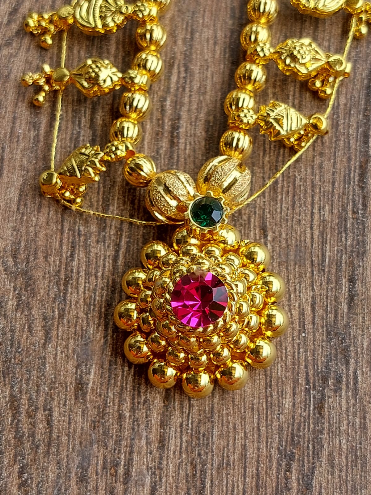 Ornate gold-toned necklace with a large floral pendant featuring a vibrant pink gemstone center and smaller green gemstone accents, displayed against a rustic wooden background.