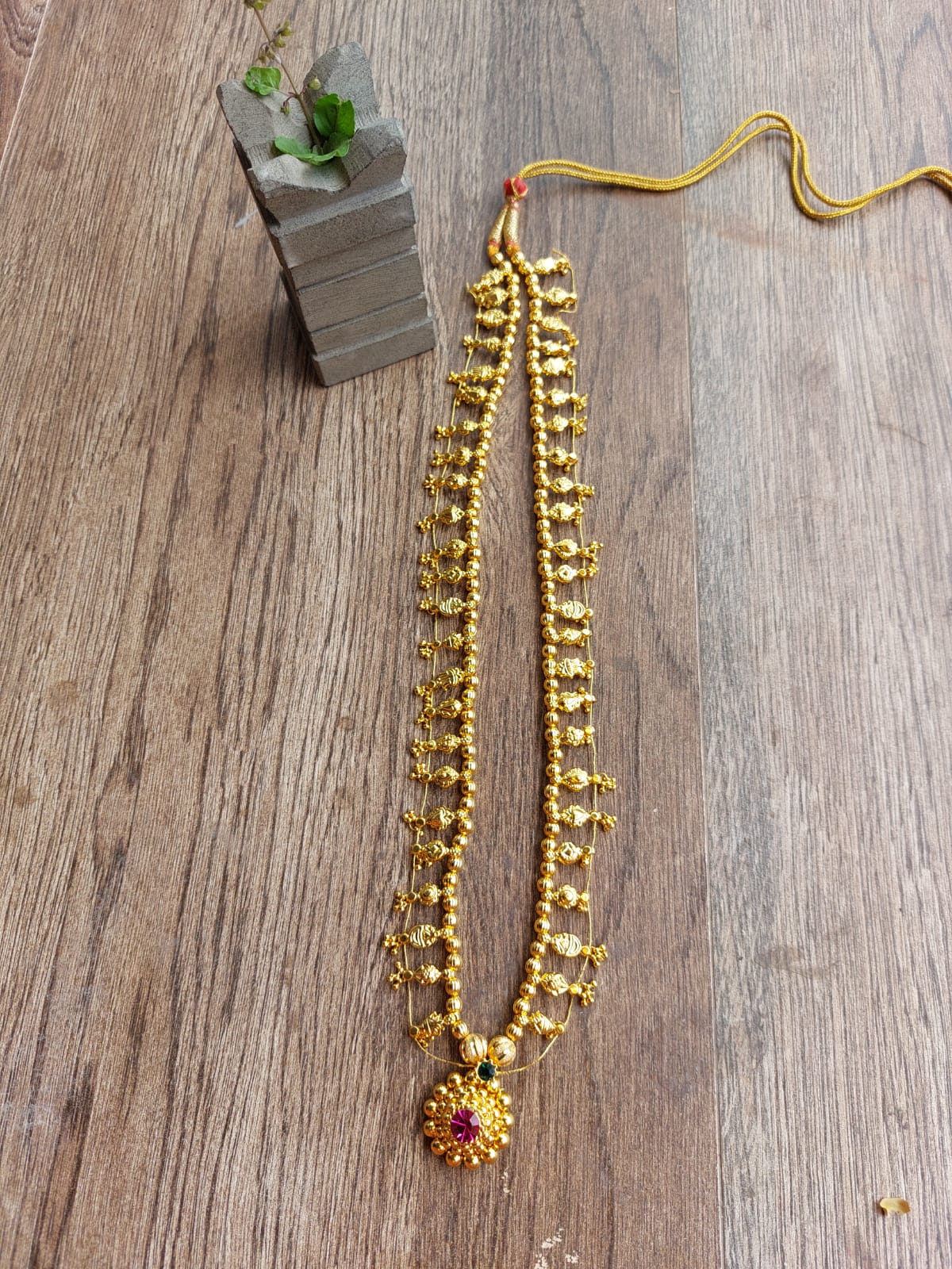 Elegant gold beaded necklace with a vibrant pendant, displayed on a rustic wooden surface with a plant accent.