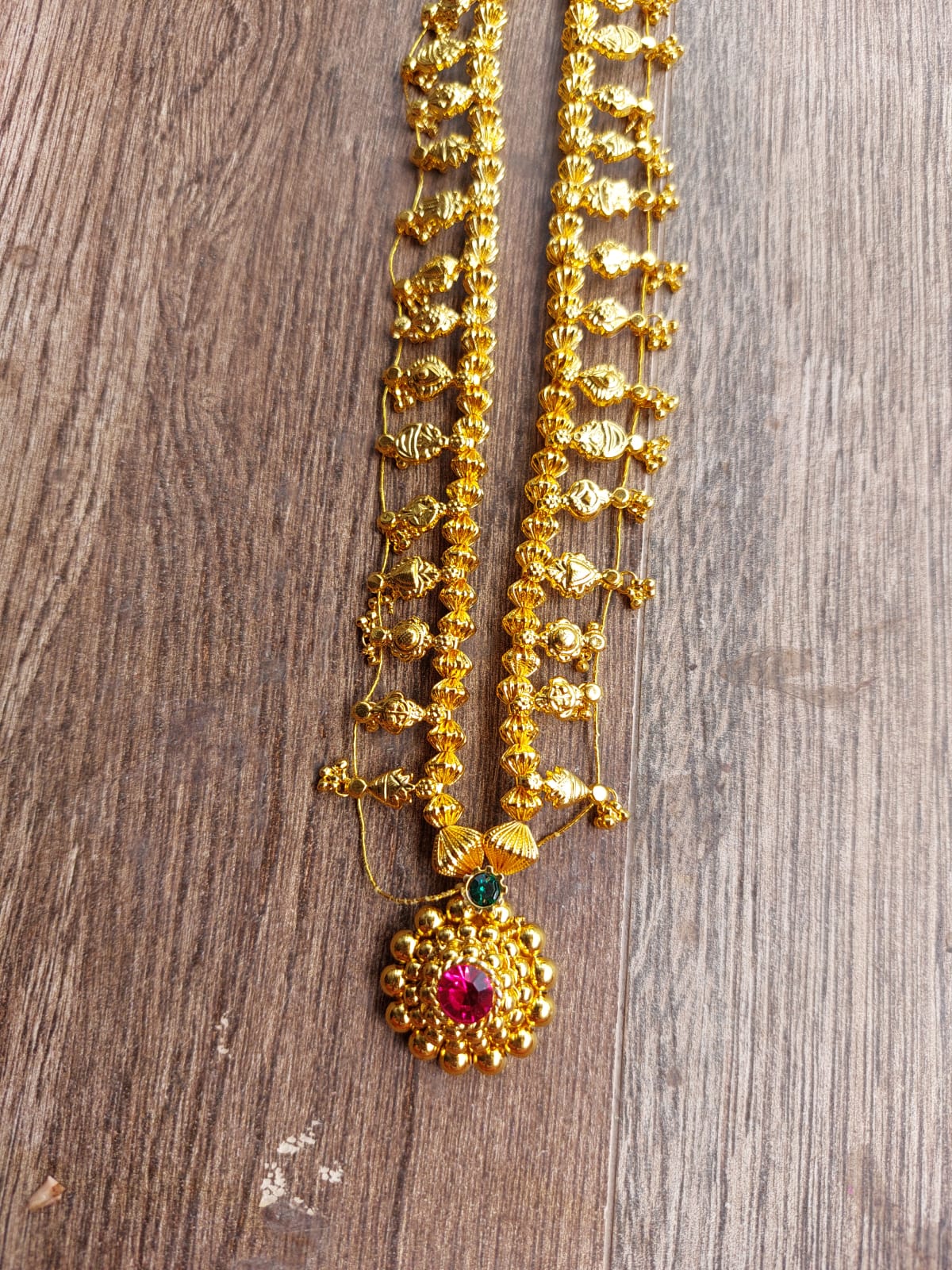 Ornate gold-toned beaded necklace with a decorative pendant featuring a red center stone, resting on a wooden surface.