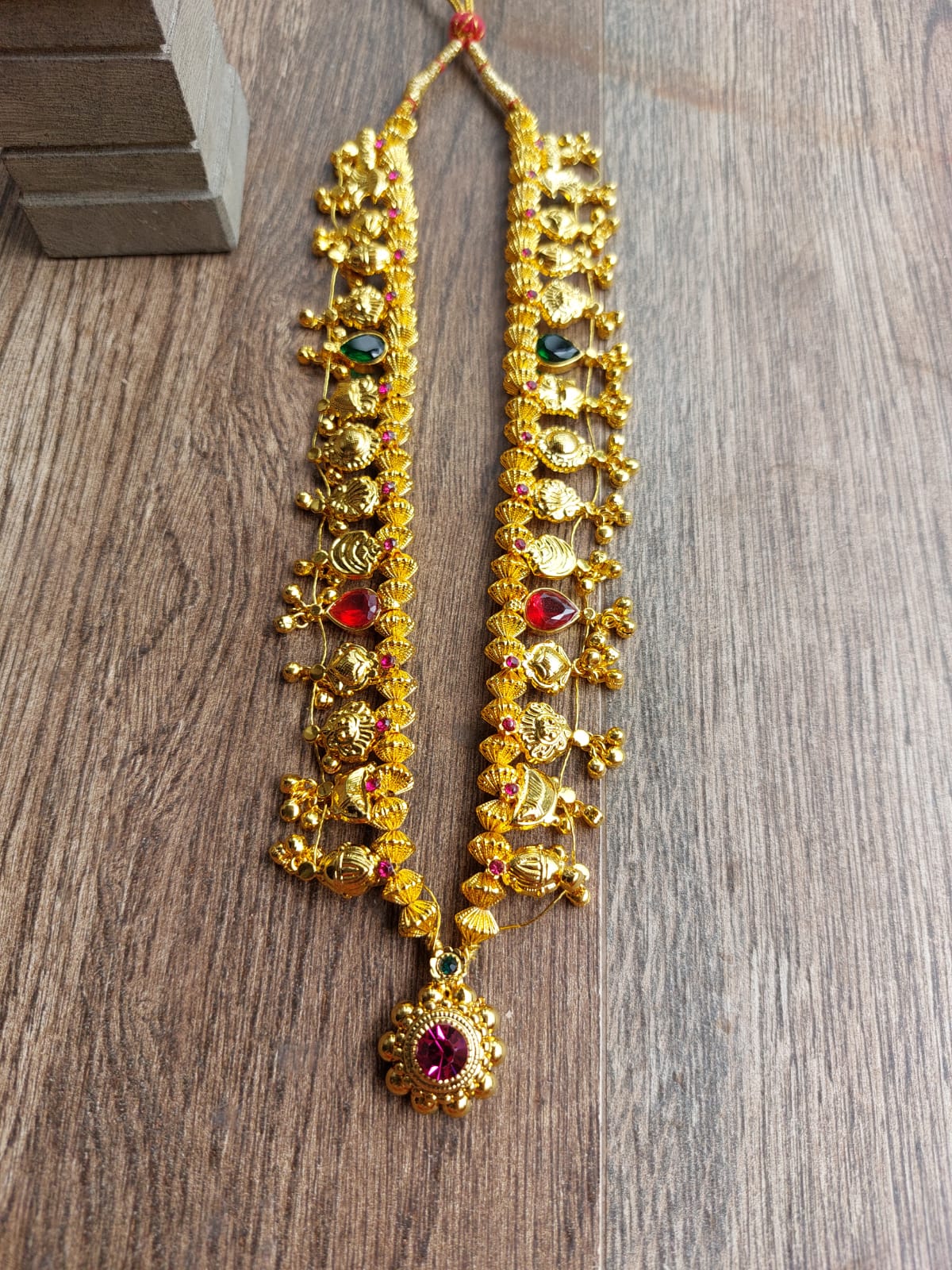 Ornate gold-colored Kolhapuri Saaj necklace with multicolored beads and gem accents, displayed on a wooden surface.