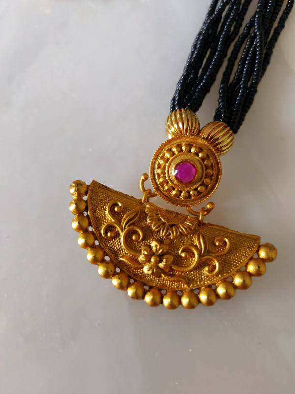 Ornate gold-tone mangalsutra pendant with intricate floral design and vibrant pink gemstone centerpiece, suspended from a long string of black beads.