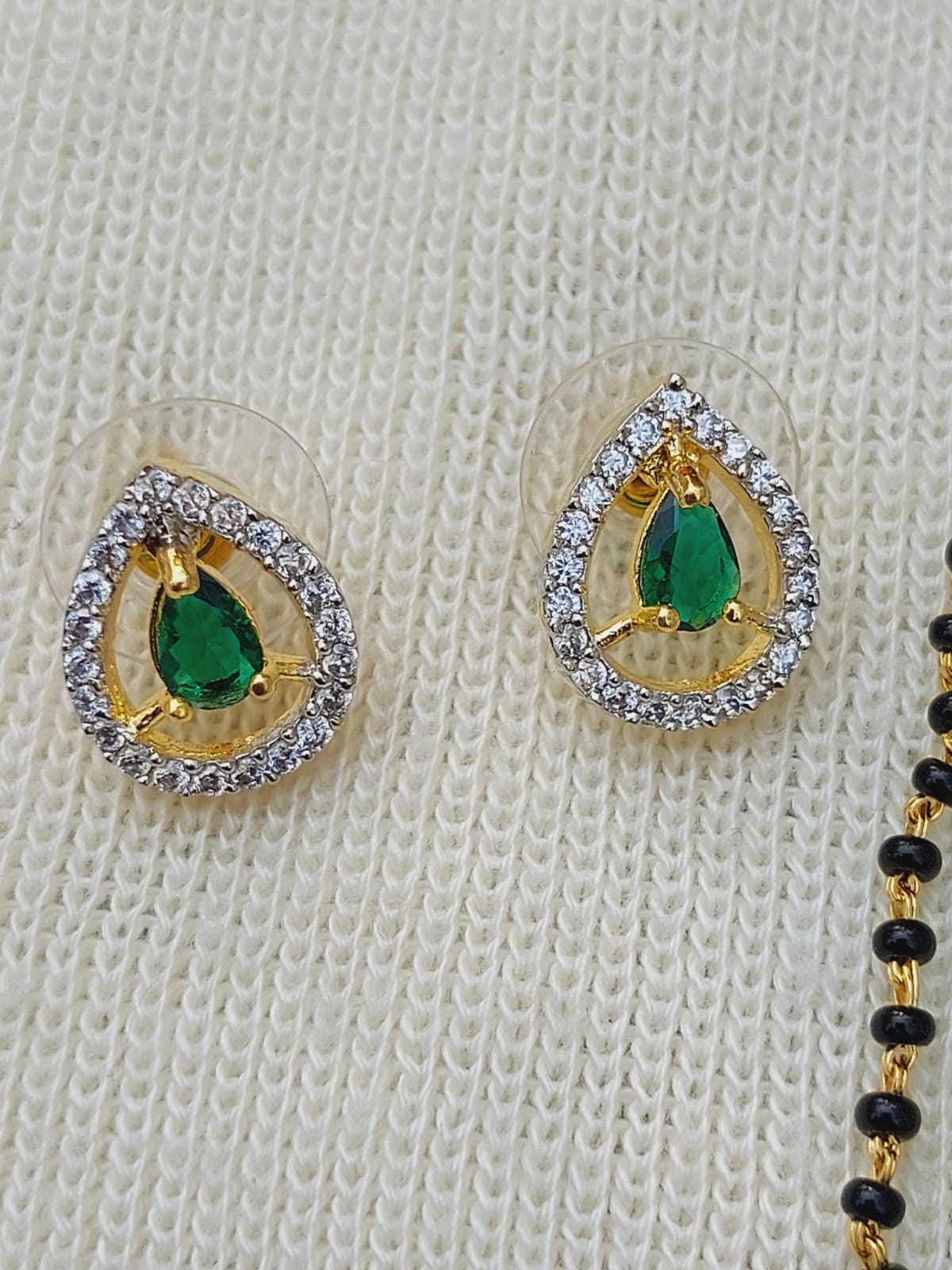 Green Stone Short Mangalsutra
Elegant gold-toned mangalsutra with delicate green stone centerpieces and sparkling diamond-like accents, set against a light-colored knit fabric background.