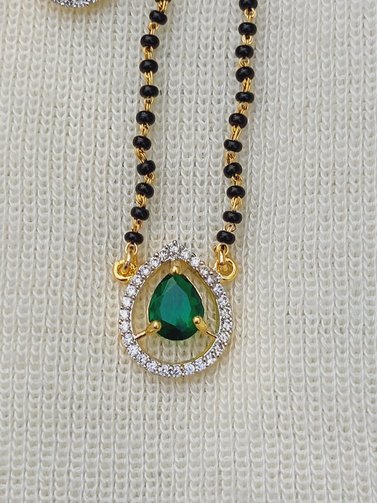 Green Stone Short Mangalsutra - Gold-tone jewelry featuring a pear-shaped green gemstone surrounded by a halo of shimmering diamonds, suspended from a beaded chain necklace.