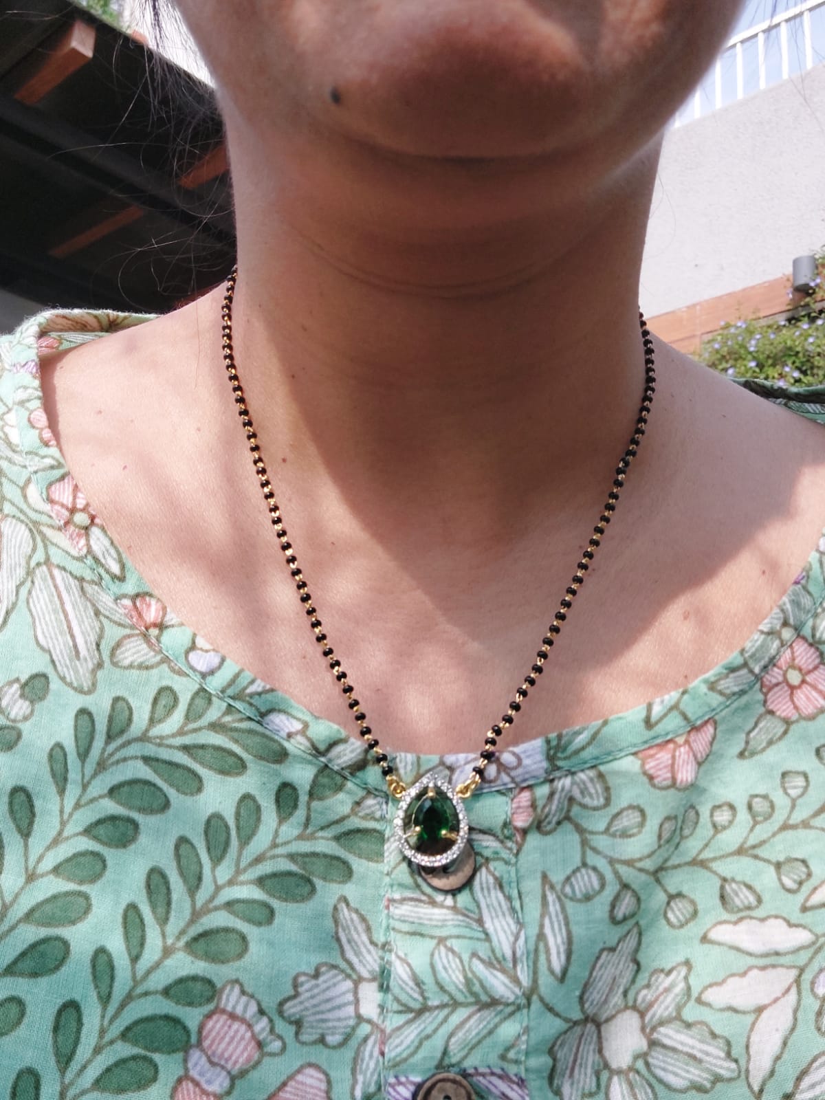 Green Stone Short Mangalsutra - Intricate necklace with green stone pendant on beaded chain, showcased on model's neck against floral patterned background.