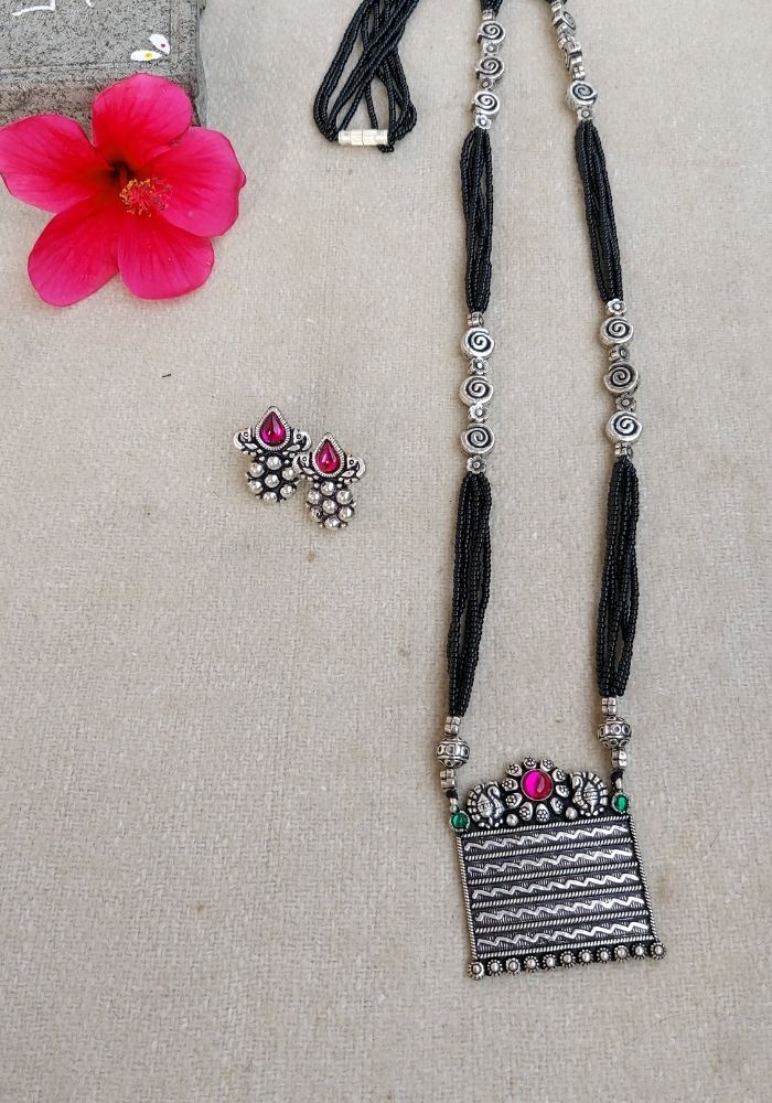 Square Pendant Designer Oxidised Long Mangalsutra
Elegant black and metallic necklace with square pendant against floral background, Mugdha Jewellery Store