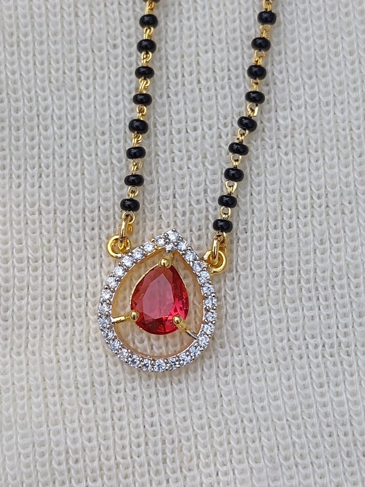 Beautiful red stone short mangalsutra necklace with delicate gold-tone chain and encrusted with sparkling stones, displayed on a white knitted background.