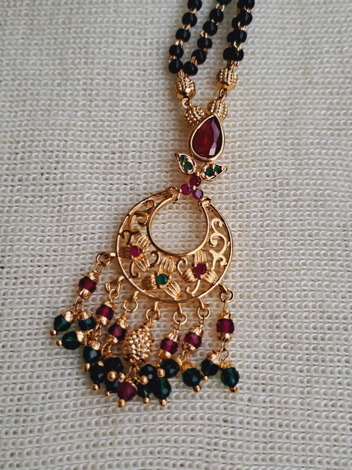 Intricate gold-toned designer pendant with dangling black beads and a vibrant red jeweled focal point, showcased on a fabric background.