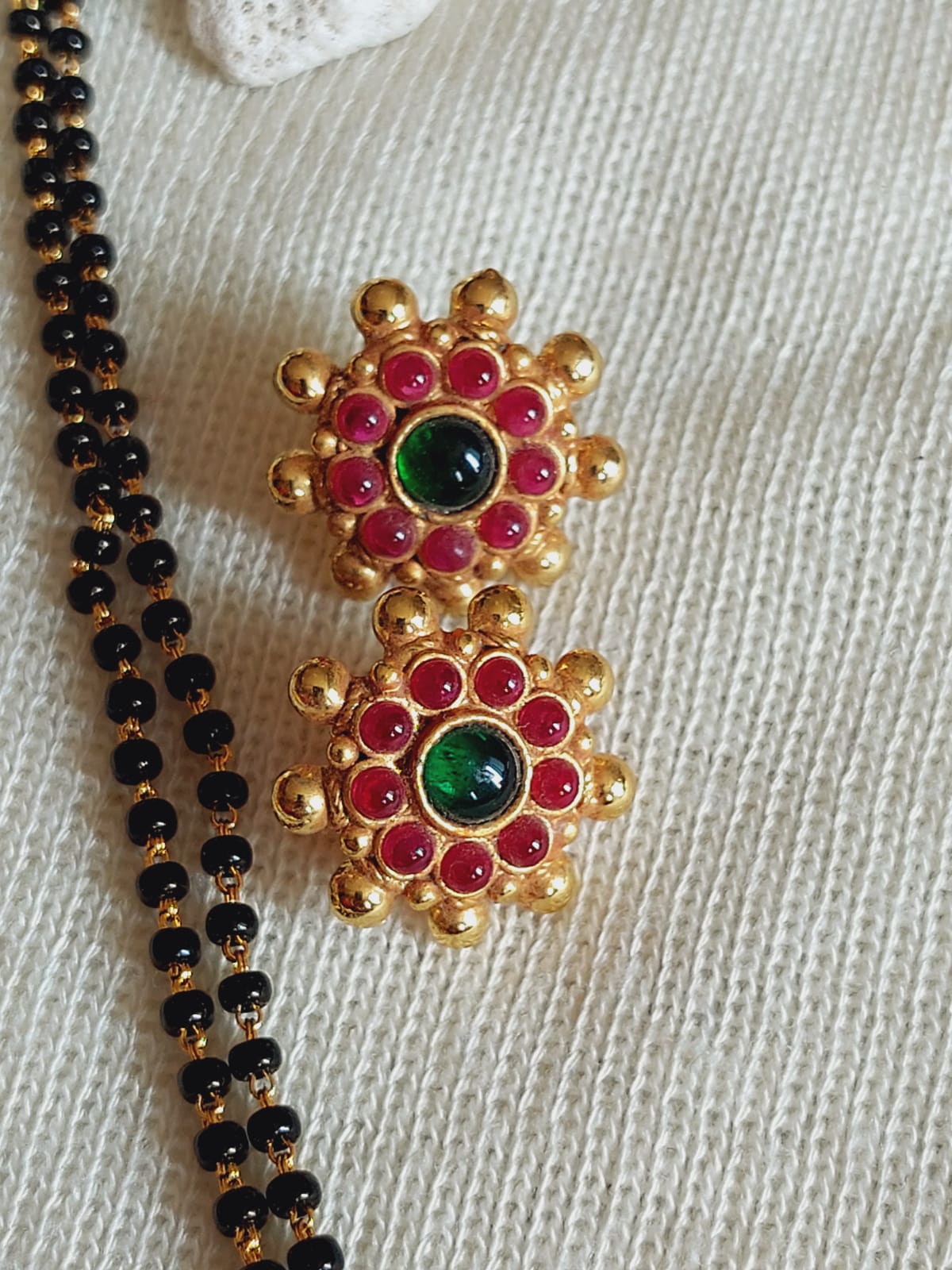 Elegant designer temple mangalsutra with golden floral accents and vibrant red and green gemstones, showcased against a soft textured background.