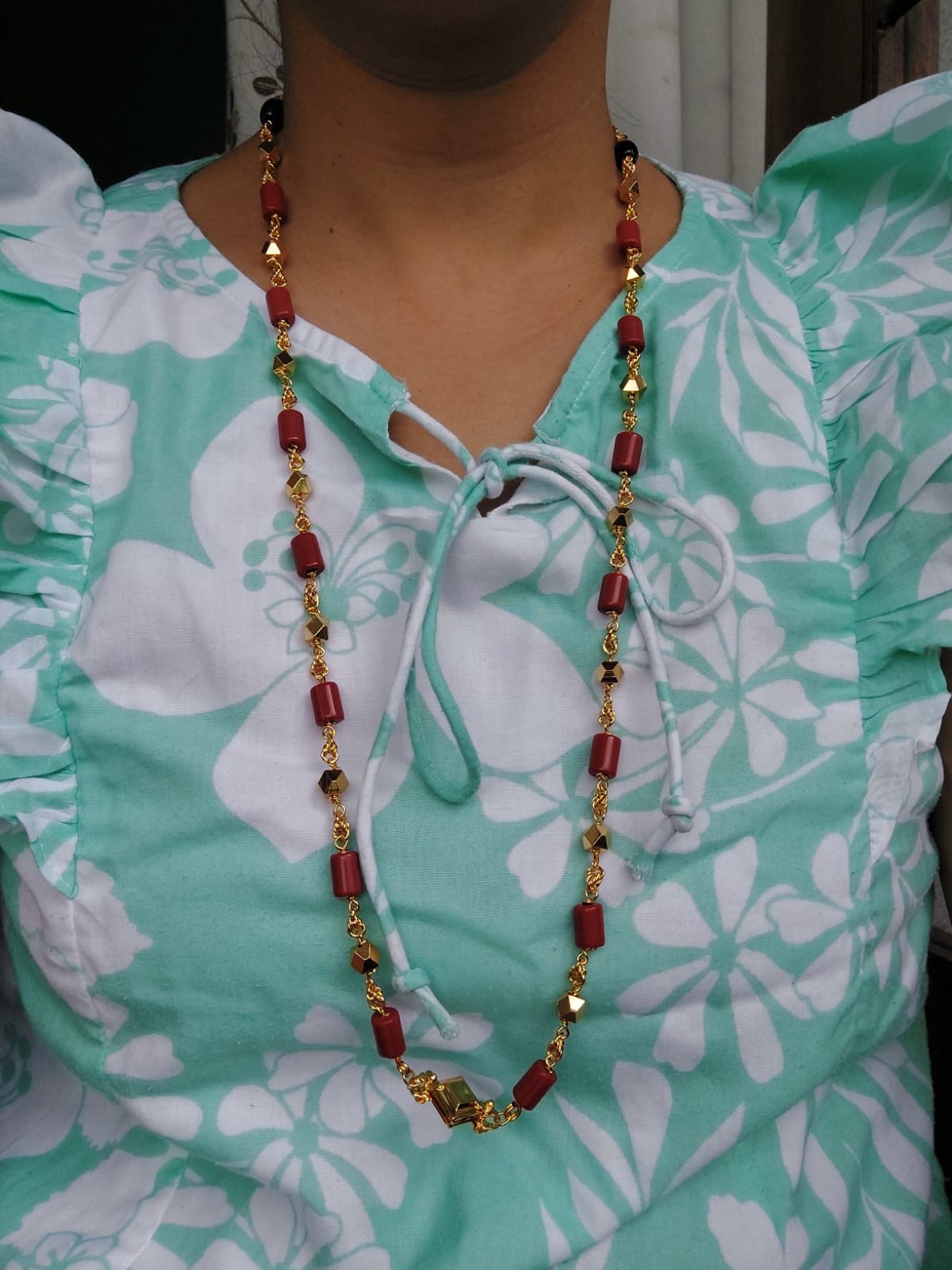 Round Beads Kashithali Long Necklace from Mugdha Jewellery Store, featuring vibrant red and gold-toned beads against a soft teal floral print blouse.