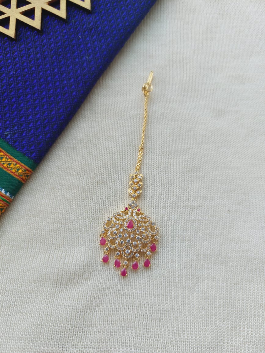 Elegant gold-colored Peacock Royal Maang Tika adorned with sparkling crystals and vibrant pink accents, showcased on a blue and white patterned background.
