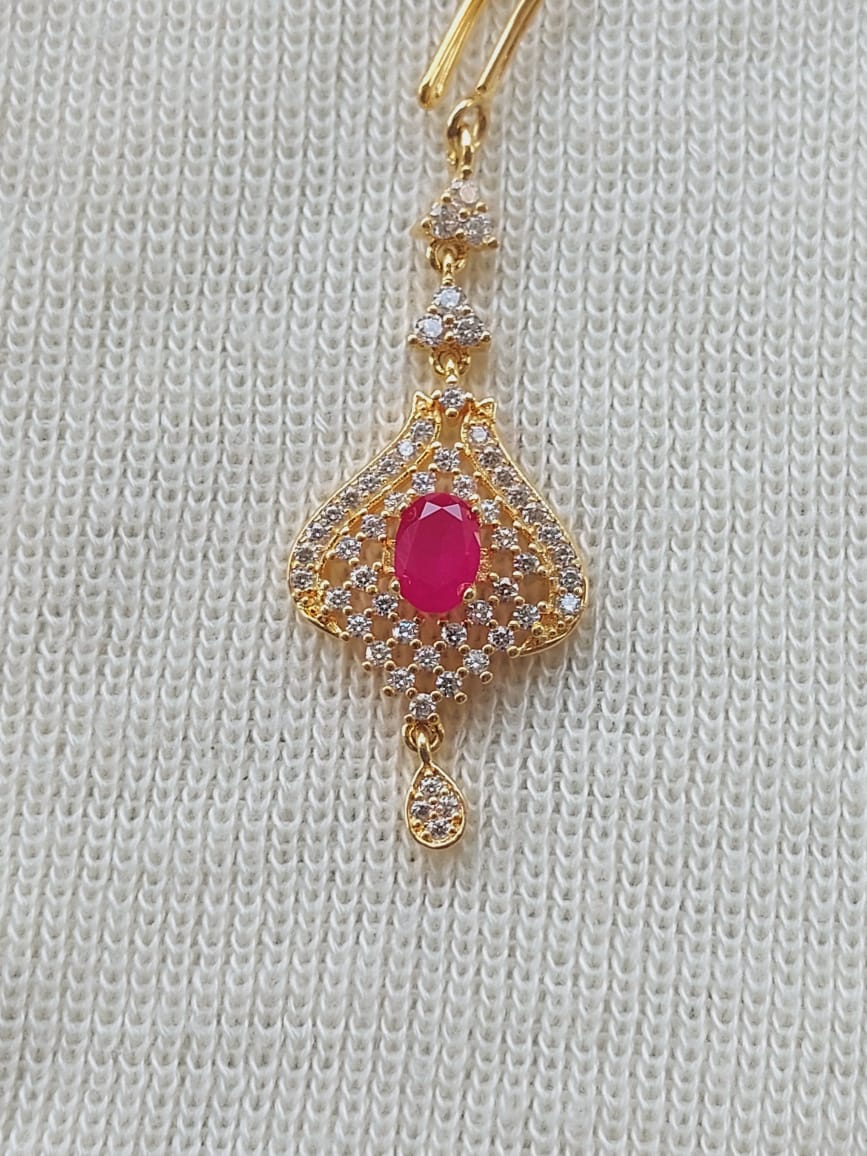 Ornate gold maang tika with a central ruby-colored gem surrounded by sparkling clear gemstones, displayed on a white fabric background.