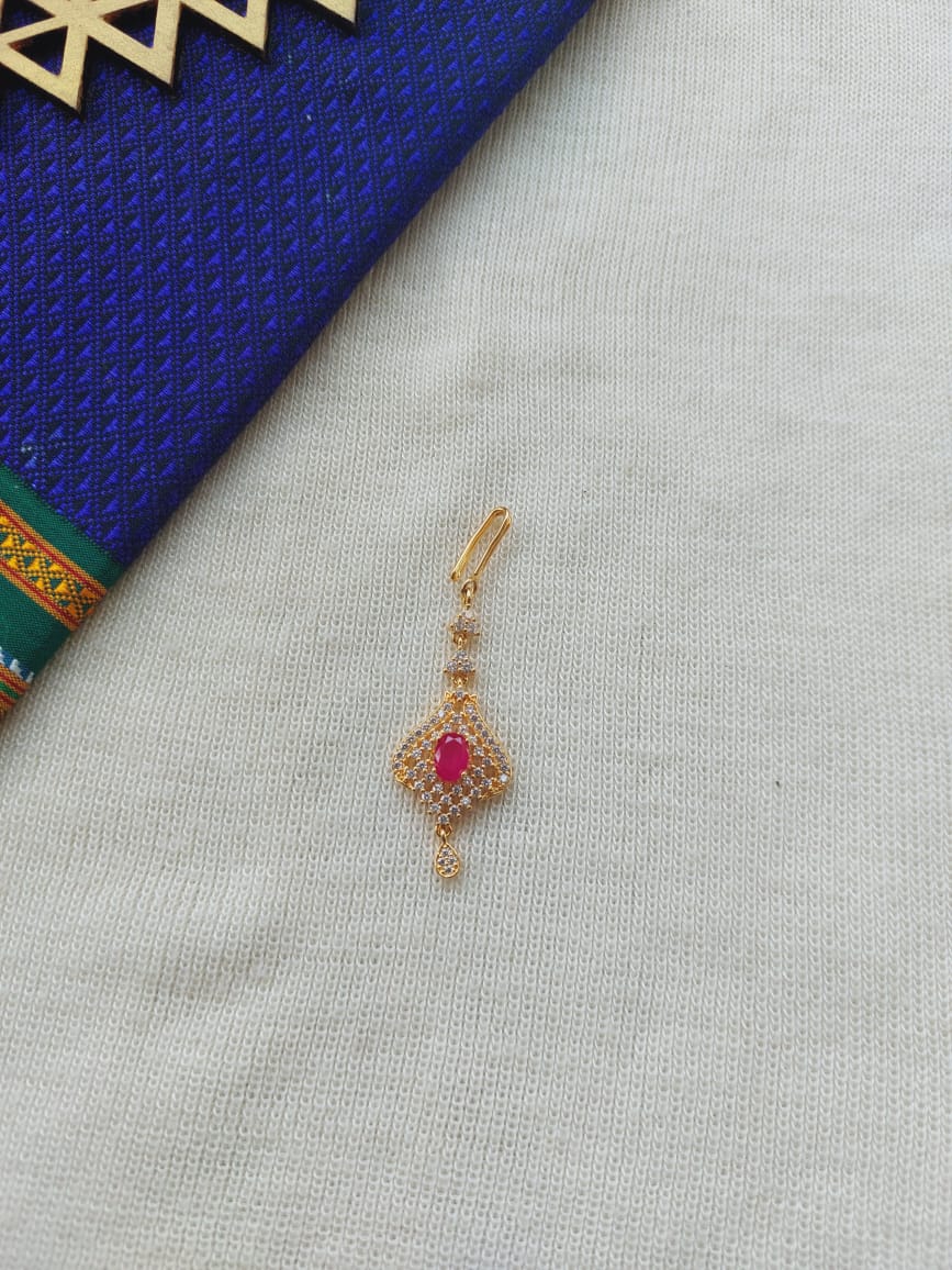 Elegant gold maang tika with a vibrant ruby center, resting on a blue and white fabric background from the Mugdha Jewellery Store.