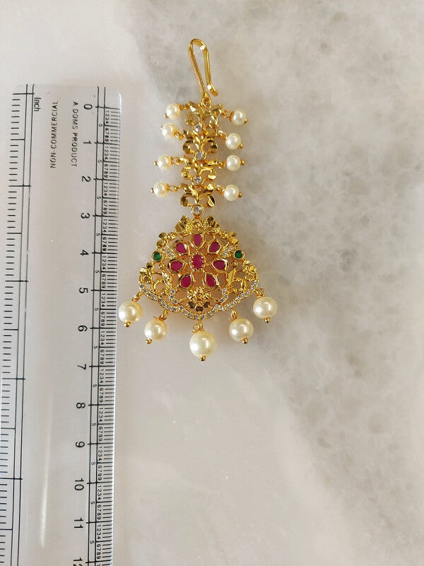 Intricate golden maang tika adorned with ruby gems and lustrous pearls, a regal accessory for the Mugdha Jewellery Store.