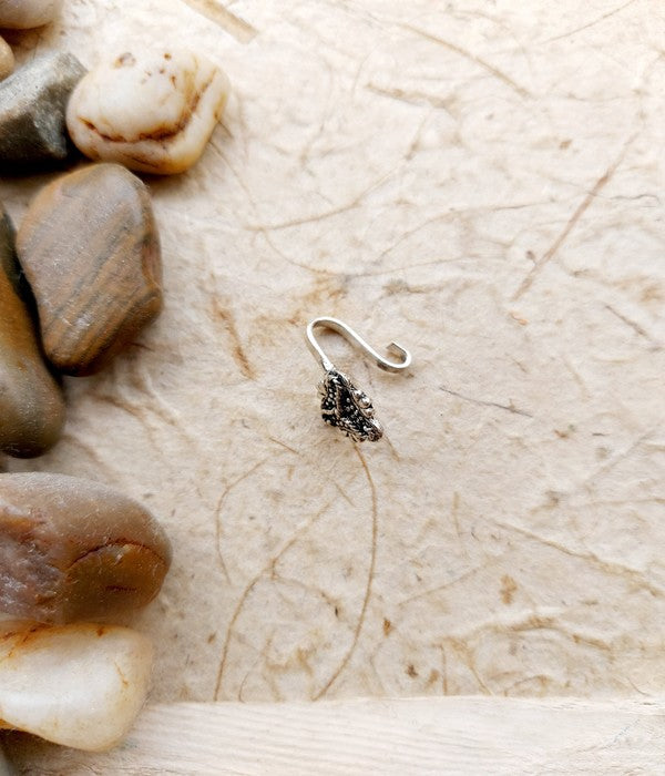Oxidised Triflower Nose Pin - Delicate metallic nose accessory with intricate floral design, surrounded by natural stones against a textured background.