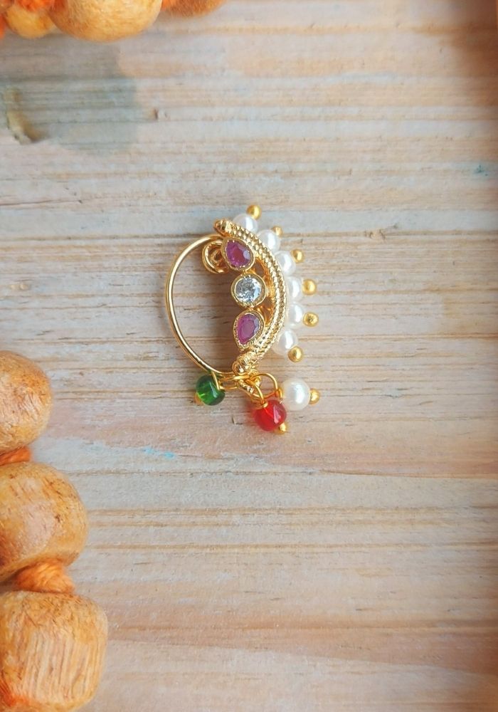 Elegant gold-toned nose ring with colorful gemstones and pearls, laid on a rustic wooden surface surrounded by orange marigold flowers.