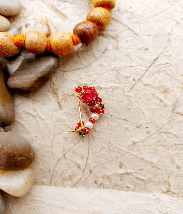 Traditional red and orange nath, a decorative nose ornament, showcased on a textured marble surface with stones and pebbles.