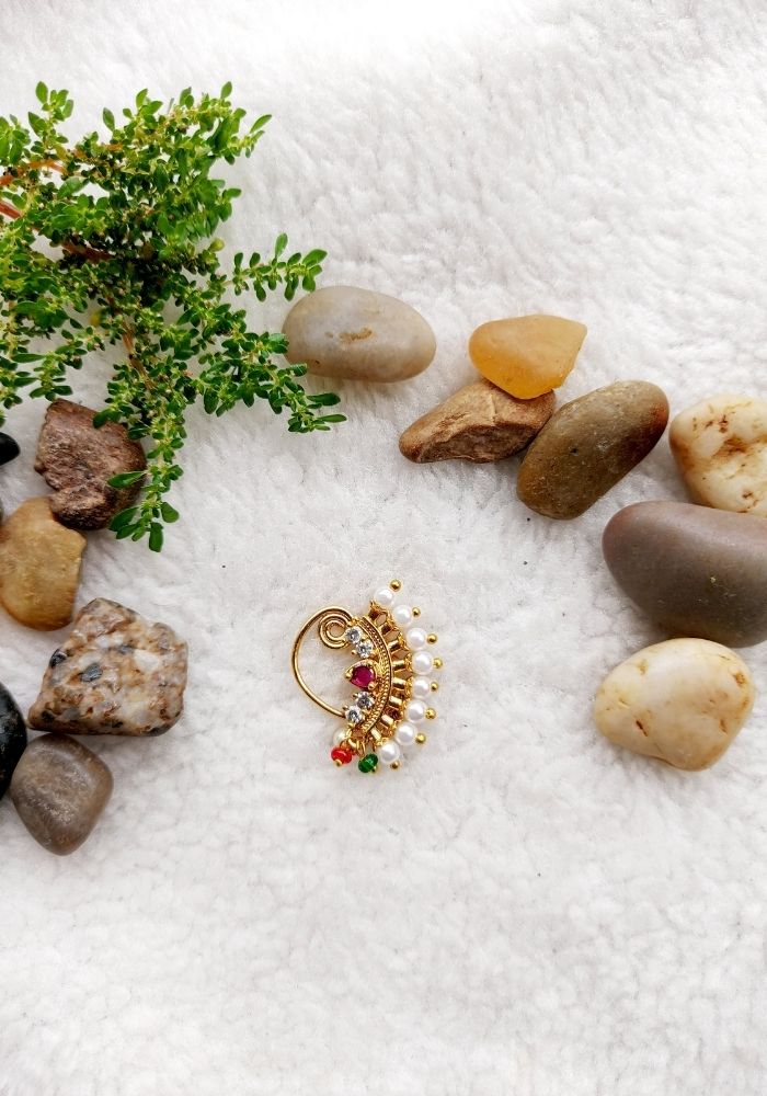 Gold jewelry with gemstones, surrounded by natural elements like greenery and smooth stones on a soft white background.