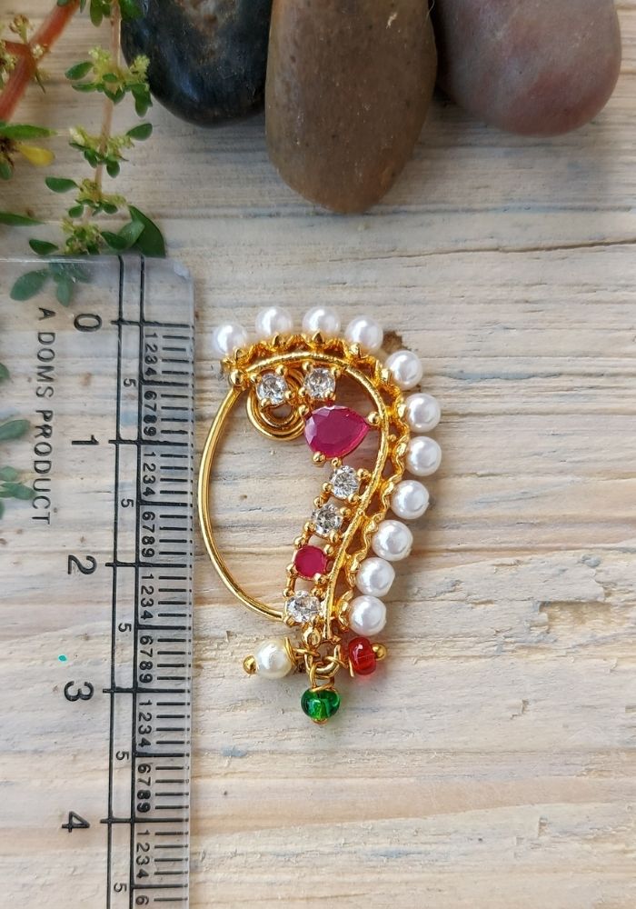 Elegant gold-toned designer Banu Nath nose pin with pearls, red gems, and sparkling rhinestones, showcased on a wooden surface alongside natural elements.