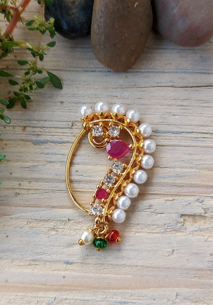 Elegant gold-toned designer Banu Nath with ornate filigree, red gemstones, and pearls, showcased on a wooden background with greenery.
