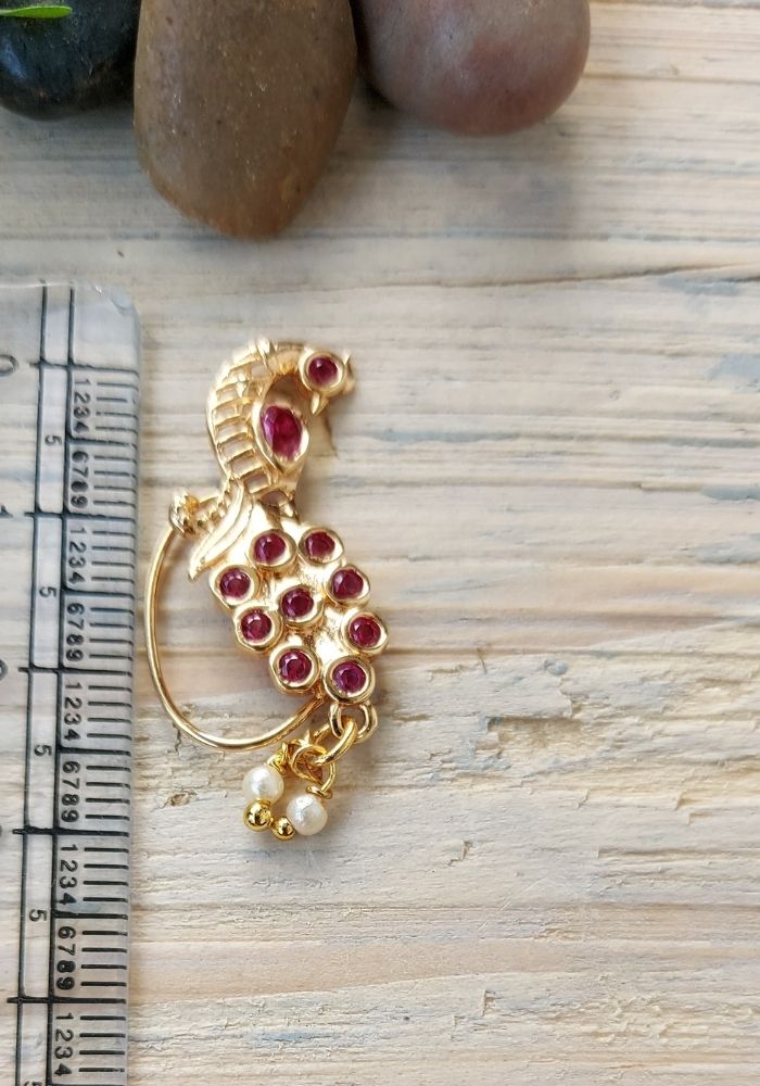 Elegant golden peacock-shaped nath adorned with vibrant red gemstones, resting on a wooden surface alongside other jewelry items.