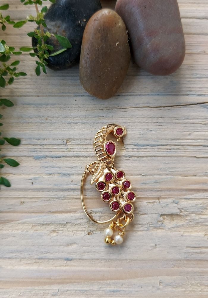 Elegant gold-toned peacock-shaped nath (nose ring) adorned with vibrant red gemstones and a dangling pearl accent, showcased on a rustic wooden surface with natural foliage elements.
