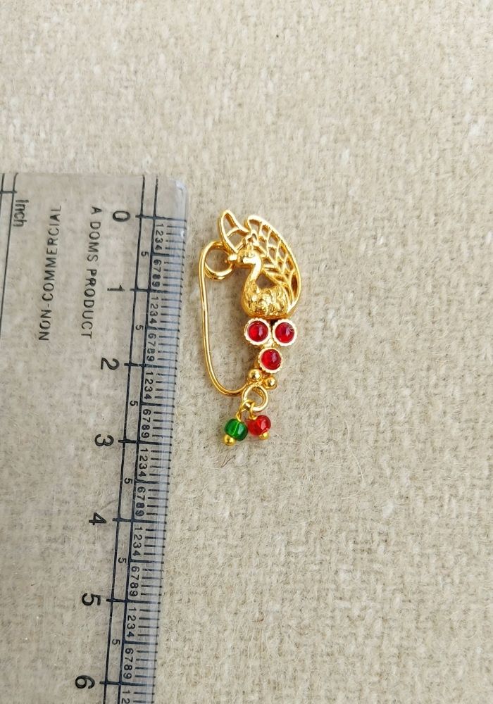 Gold-toned peacock-shaped nath with red and green gemstone accents, placed on a ruler for scale