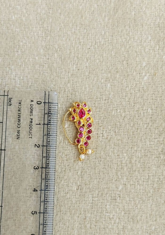 Ornate gold and ruby nath (nose ring) placed on a ruler against a plain background.