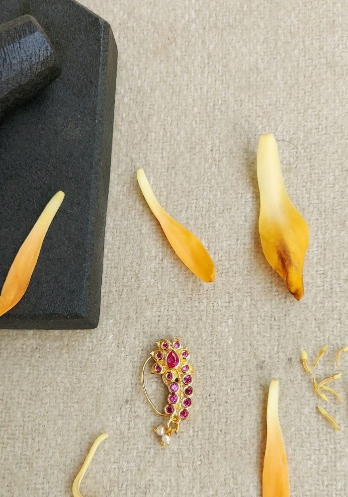 Elegant leaf-shaped gold nath with ruby accents on a gray display stand, surrounded by stylized yellow petals or leaves on a light background.