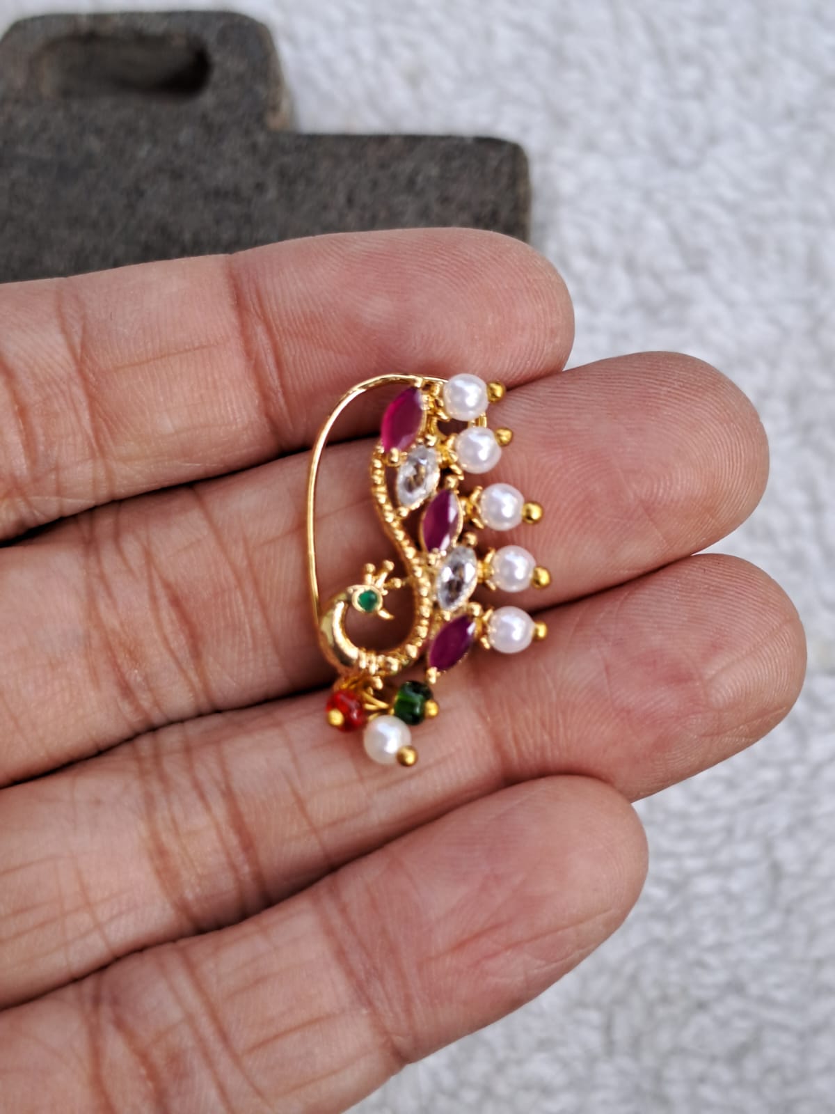 Ornate gold-toned nath with vibrant ruby gemstones, pearls, and green accents, showcased on a hand against a plain background.