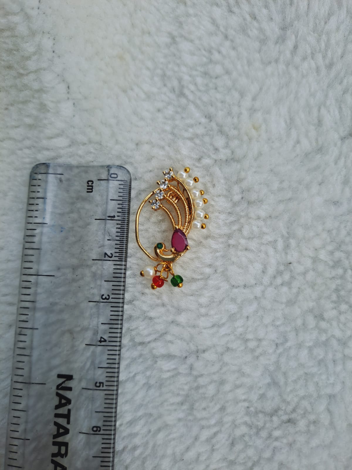 Elegant gold-toned Dancing Peacock Nath with vibrant ruby and pearl accents, placed on a ruler against a soft white background.