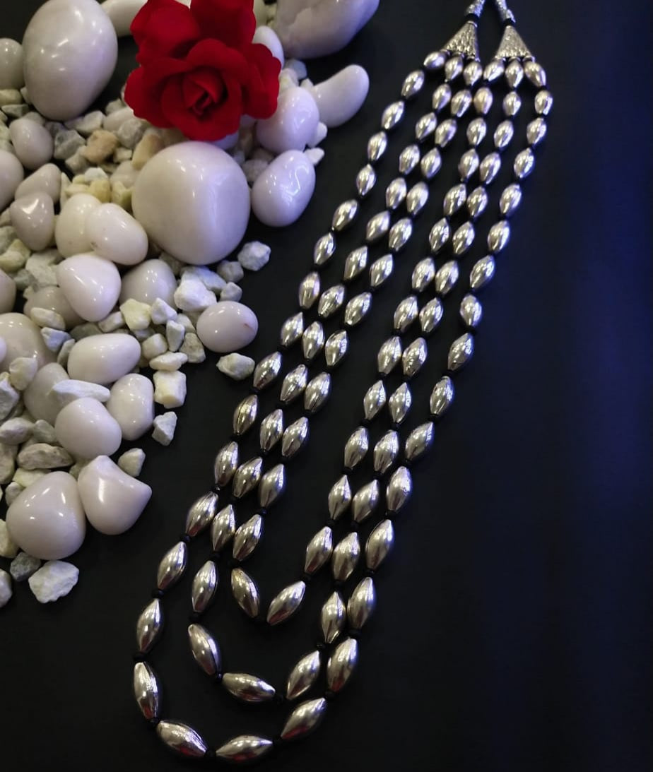 Oxidized long necklace with 3 lyre dholki beads, surrounded by white stones and a red rose on a dark blue background.