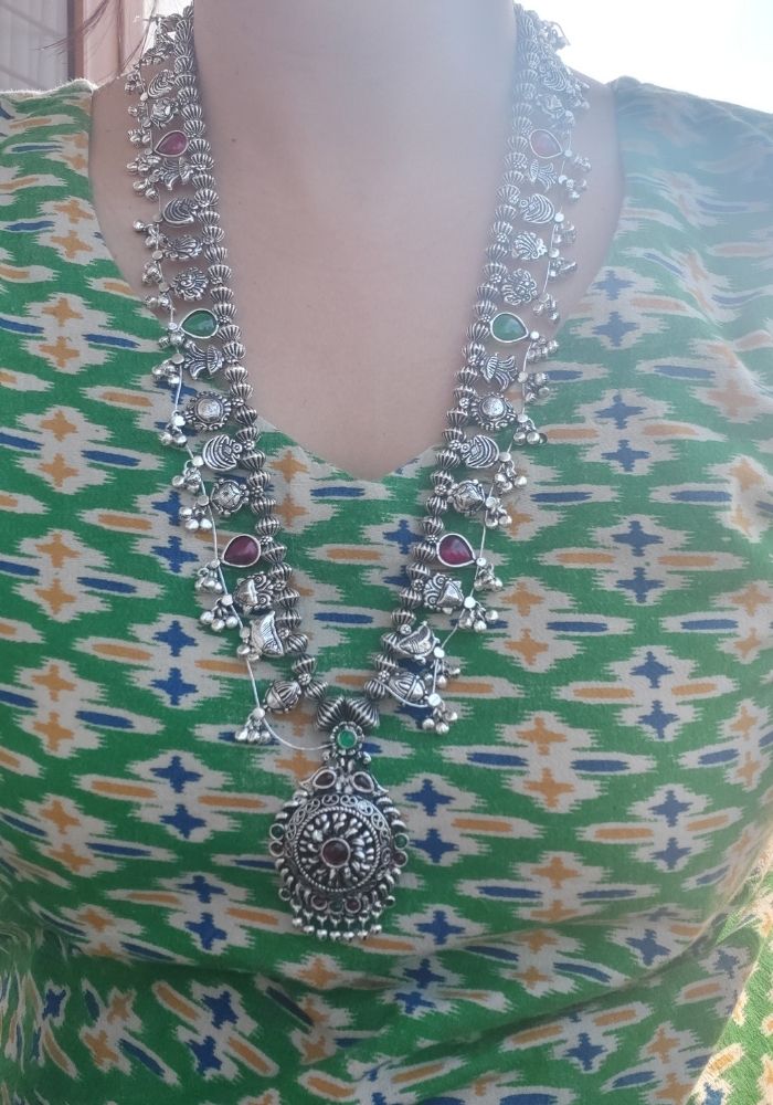 Ornate silver-toned necklace with intricate floral and geometric designs, featuring a large pendant with a circular pattern. The necklace is placed on a green and white patterned textile, adding a vibrant and ethnic touch to the image.