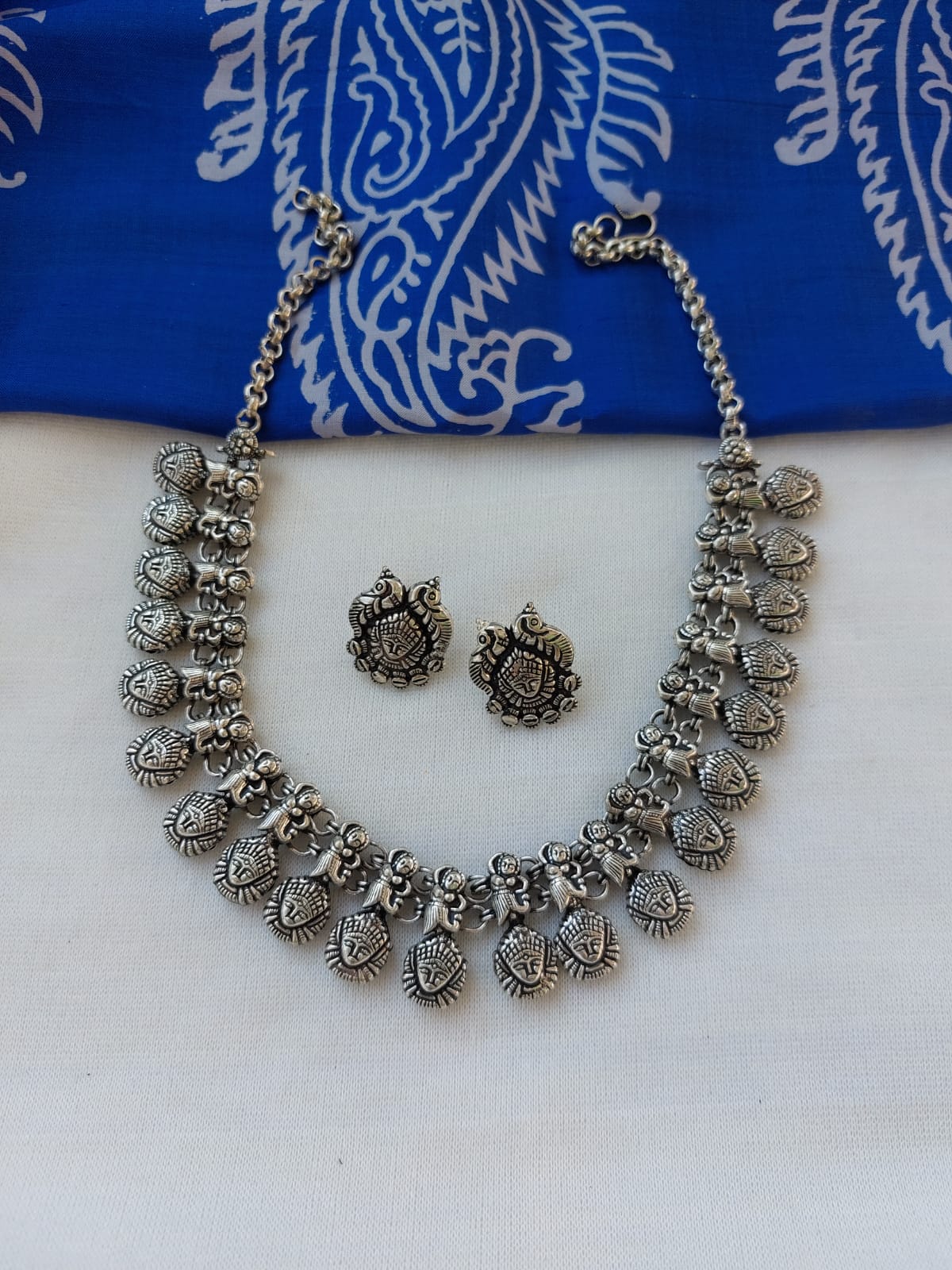 Ornate oxidised silver necklace set displayed on a vibrant blue patterned backdrop, featuring intricate pendants and studs in a traditional Indian design.