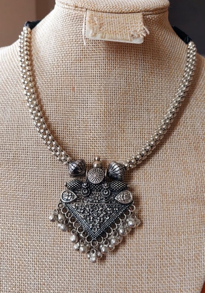 Elegant oxidized metal necklace with intricate floral pendant adorned with studded crystals, displayed on a neutral-toned display stand in the Mugdha Jewellery Store.