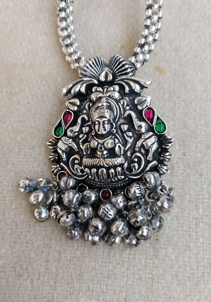 Ornate silver-toned pendant featuring a detailed Hindu deity figure surrounded by colorful gemstones and metal beads, displayed on a multi-strand chain necklace from the Mugdha Jewellery Store.