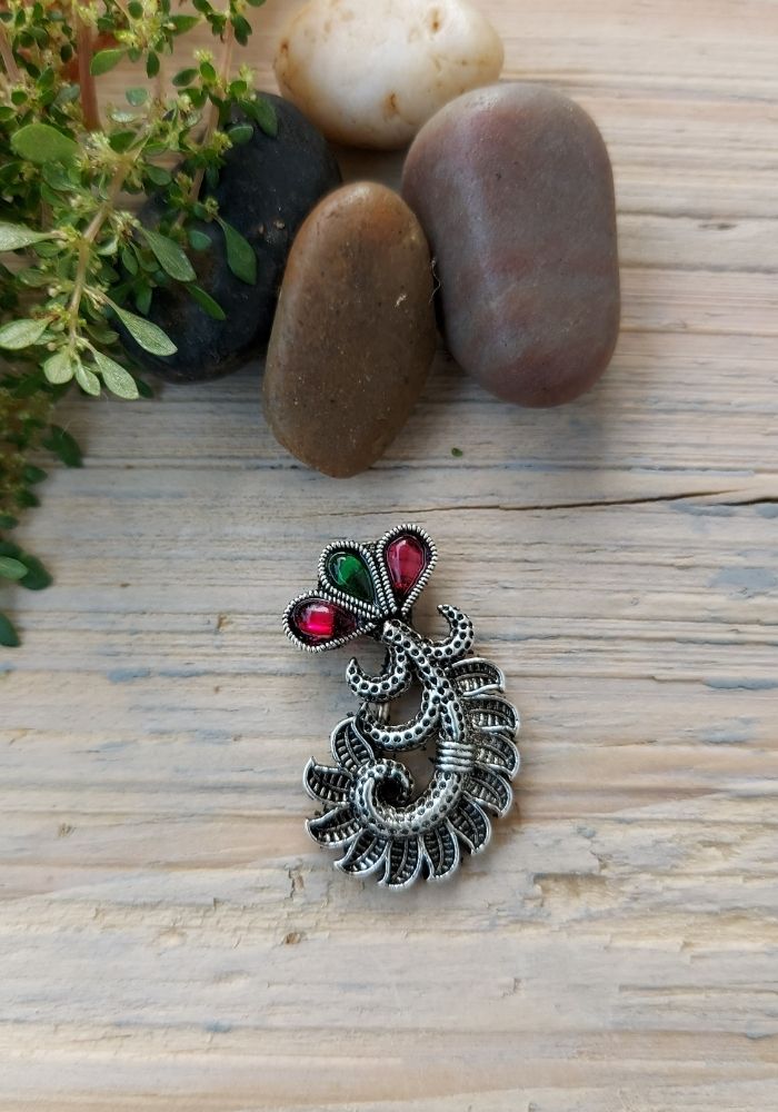 Ornate oxidized silver brooch with colorful gemstone accents, placed on a rustic wooden surface alongside fresh herbs and vegetables.
